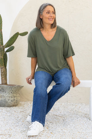 Bamboo V-Neck T-Shirt in Moss, full length view on model seated outdoors, styled with denim jeans and white sneakers.