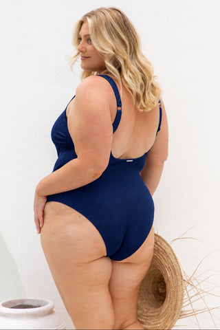 A woman modeling a navy blue ribbed one-piece swimsuit from the back, standing against a white wall with a straw hat and a white pot nearby.