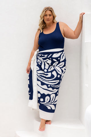 A woman modeling a navy blue and white patterned sarong wrap tied around her waist, paired with a solid navy blue tank top, standing against a white wall.